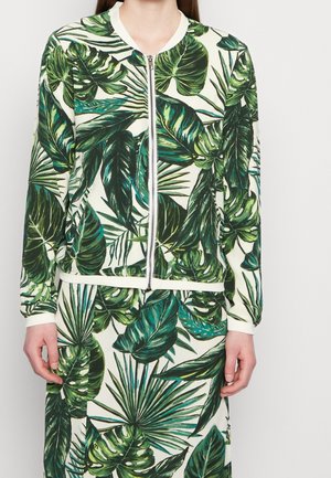 Vrouw draagt een jas met rits en rok met een groen tropisch bladerenpatroon op een witte achtergrond.