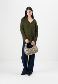 Grønn v-hals genser, blå vide bukser, holder en beige veske med slangemønster i en buet form og teksturert overflate.