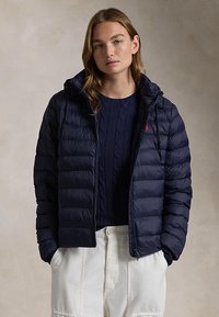 Frau trägt eine marineblaue gesteppte Jacke über einem marineblauen Zopfmuster-Pullover und weißen Hosen, steht mit den Händen in den Taschen vor neutralem Hintergrund.