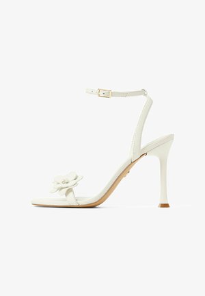 Sándalo blanco de tacón alto con tira en el tobillo, que presenta un detalle de flor en la punta y un acabado suave, montado sobre un delgado tacón de aguja.