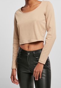 Beiges, langärmliges, cropped Oberteil aus weichem Stoff mit einem runden Ausschnitt. Kombiniert mit hochgeschnittenen schwarzen Lederimitathosen.