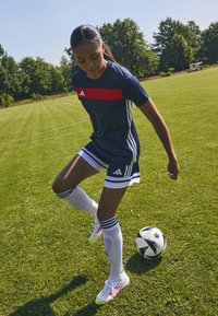 Maillot de sport Adidas bleu marine et rouge avec des rayures blanches, associé à des chaussettes blanches et des crampons, porté tout en dribblant un ballon de soccer sur l'herbe.