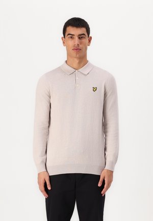 Camisa polo de manga larga color beige claro con una textura suave, cuello y un pequeño logo de mariposa amarilla en el lado izquierdo del pecho. Pantalones negros.