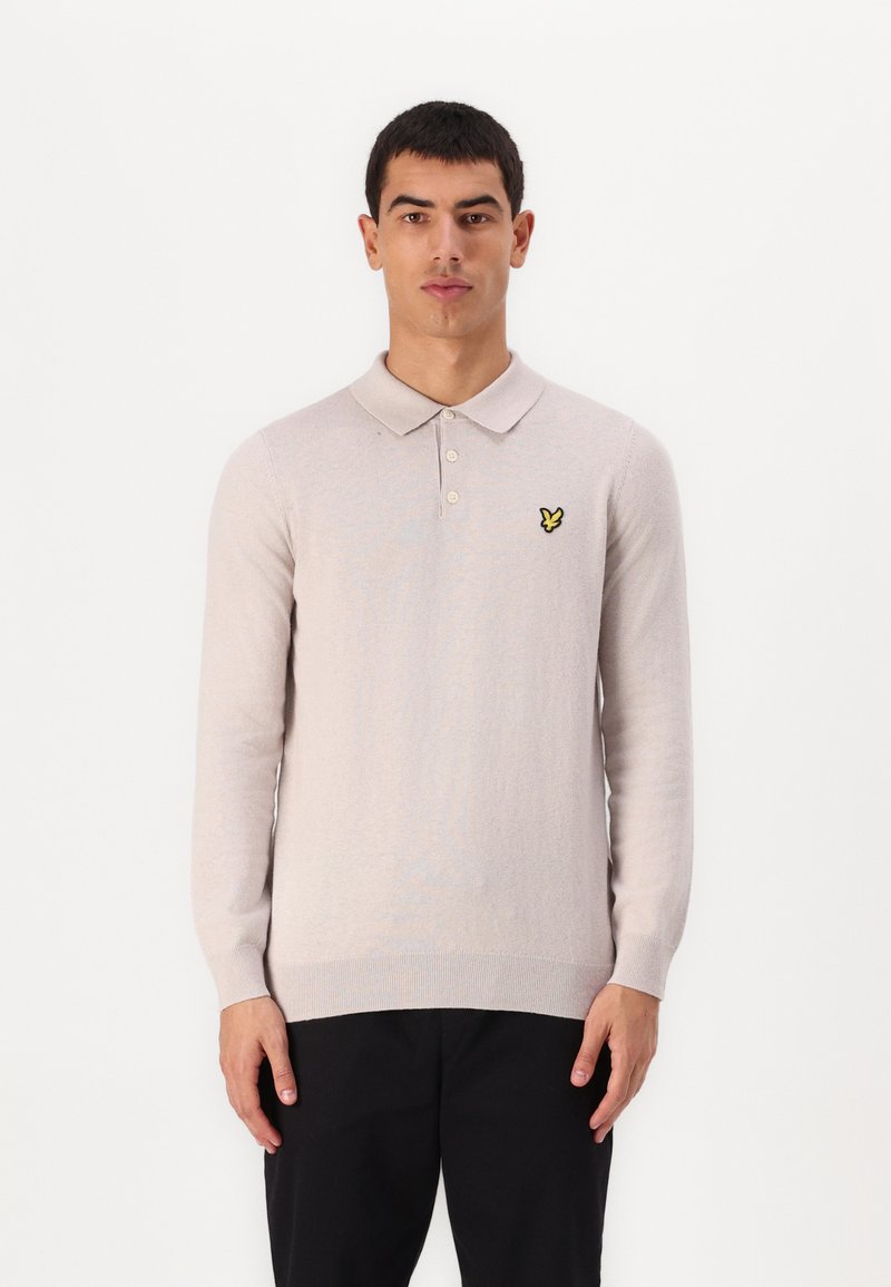 Polo shirt a maniche lunghe color beige chiaro con una texture morbida, colletto e un piccolo logo a forma di farfalla gialla sul lato sinistro del petto. Pantaloni neri.