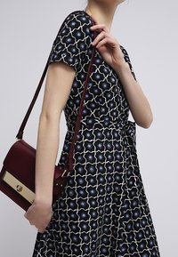 Kvinna i en svart mönstrad klänning med blå och vita blommor, hållande en vinröd axelväska med ett guldspänne.