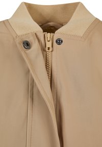 Giacca beige con colletto testurizzato, chiusura a scatto in metallo e zip. Presenta una superficie di tessuto liscia e dettagli di cucitura puliti.