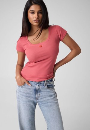 Jeune femme aux longs cheveux foncés portant un haut corail ajusté à col rond et manches courtes, ainsi qu'un jean taille haute bleu clair, posant avec une main dans la poche.