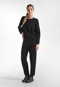 Zwarte gebreide trui met lange mouwen, geribbelde manchetten en tailleband, gecombineerd met zwarte broeken. Model draagt zwarte sneakers. Glad textuur.