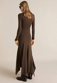 Robe marron à manches longues présentant un ourlet fluide et asymétrique, en tissu texturé. Associée à des bottines noires et des bracelets superposés.