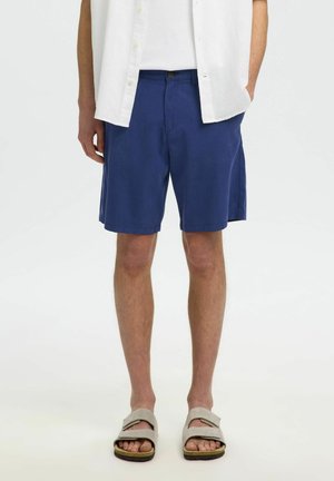 Homme portant un short bleu jusqu'aux genoux, une chemise blanche boutonnée et des sandales beige, debout devant un fond uni.