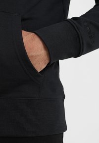 Sudadera negra con puños y dobladillo acanalados, y un bolsillo tipo canguro. El material parece suave y texturizado. Detalle sutil del logo en la manga.