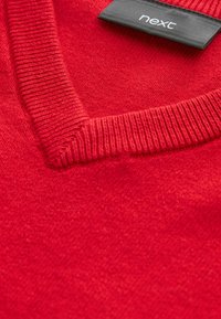 Gros plan sur un pull en maille rouge à col en V avec une étiquette en tissu noir portant la mention "next" cousue à l'intérieur du col.