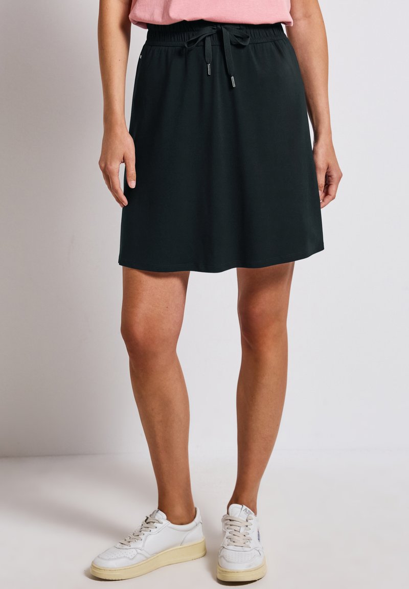 Jupe noire à enfiler avec une taille élastique et un cordon de serrage ajustable. Longueur genou avec une texture lisse, associée à des baskets blanches.