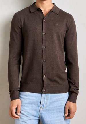 Man draagt een bruine cardigan met knopen, lange mouwen, kraag en lichtblauwe spijkerbroek tegen een effen achtergrond.