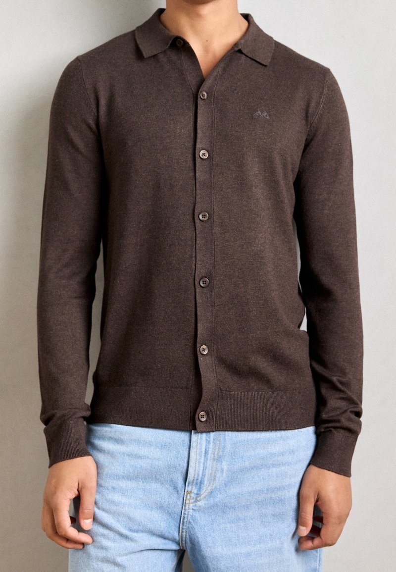 Homme portant un cardigan marron boutonné à manches longues avec un col et un jean bleu clair sur un fond uni.