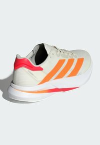 Zapatilla de running ligera en malla color blanco roto con acentos en naranja brillante y rosa, que presenta una suela blanca acolchada y un collar acolchado.