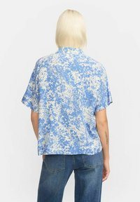 Short-sleeve, collared shirt in blue with white splattered pattern, made from lightweight fabric. Worn with high-waisted denim jeans.