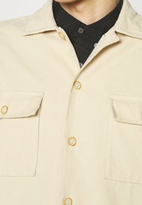 Giacca in corduroy beige chiaro con colletto, bottoni gialli, due tasche frontali e tessuto strutturato. La camicia interna è nera con bottoni.