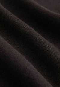 Detailní záběr na měkkou, tmavě hnědou látku s viditelnou pletenou texturou a jemnými záhyby, které vytvářejí stín a hloubku.