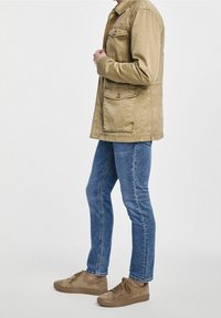 Homme portant une veste beige avec plusieurs poches, un jean slim bleu et des baskets en daim beige, debout devant un fond uni.