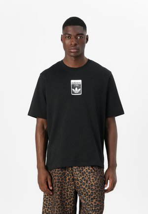 Black cotton t-shirt featuring a white Adidas logo patch at the chest. Model is wearing leopard-print shorts. Short sleeves, round neckline.