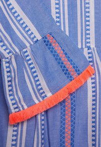 Tela azul a rayas con patrones horizontales y verticales, con un borde con flecos en un intenso color coral. Textura suave, detalles del diseño tejida visibles.