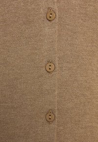 Beige neulottu neulejakku lähikuvassa, jossa on viisi ruskeaa nappia edessä, sileä pinta ja hienovarainen ribbineuloskuvio.