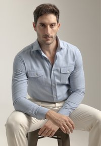 Camisa de manga larga azul claro hecha de tela texturizada, con dos bolsillos en el pecho y botones. Combinada con pantalones de color crema.