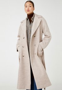 Helle beige, doppelt modische Jacke mit großen Revers, Seitentaschen und metallischen Knöpfen; über einem karierten Hemd getragen. Glatte Textur.