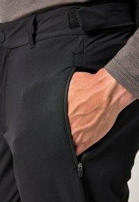 Pantalones deportivos negros con un bolsillo lateral con cremallera y cierre de Velcro, que presentan una textura suave y un corte ajustado. Mano descansando en el bolsillo.