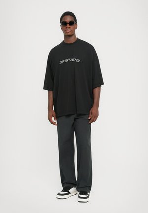 Young man wearing black oversized t-shirt with white lettering, loose dark pants, black sunglasses, and black-and-white sneakers, standing straight.