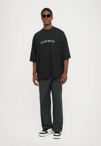 Jonge man in een zwart oversized t-shirt met witte letters, losse donkere broek, zwarte zonnebril en zwart-witte sneakers, staat rechtop.