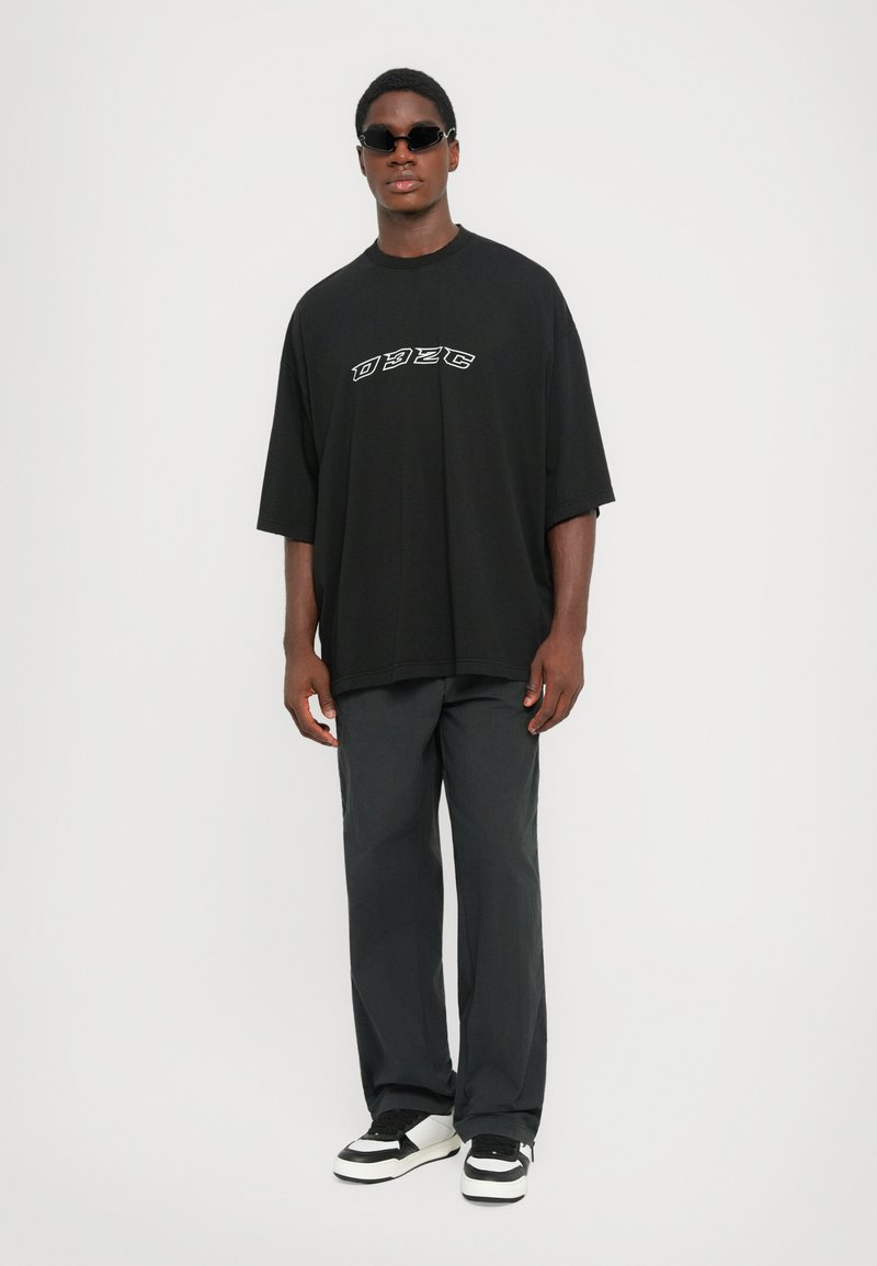 Young man wearing black oversized t-shirt with white lettering, loose dark pants, black sunglasses, and black-and-white sneakers, standing straight.
