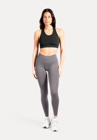 Reggiseno sportivo nero con design aderente, abbinato a leggings grigi a vita alta. Modello con scarpe da ginnastica bianche, in piedi su uno sfondo neutro.