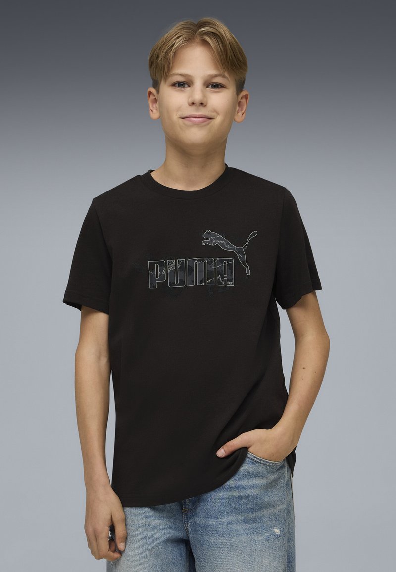 Šviesiaplaukis paauglys vaikinas, vilkintis juodą „Puma“ marškinėlį ir šviesiai mėlynas džinsas, stovintis su viena ranka kišenėje prieš pilką gradientinę foną.