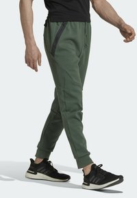 Zelené joggingové kalhoty vyrobené z měkké tkaniny, s černými kapsami na zip, zúženým střihem a elastickými manžetami, doplněné černými sportovními botami.