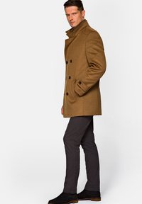 Brązowy, podwójnie zapinany płaszcz z wełny, z czarnymi guzikami, kieszeniami po bokach i wysokim kołnierzem, w połączeniu z ciemnoszarymi spodniami w fasonie slim fit i czarnymi botkami.