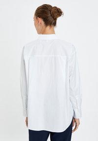 Camisa blanca de manga larga con cuello clásico, detalles de botones y un corte relajado. Textura suave con un dobladillo ligeramente curvado.
