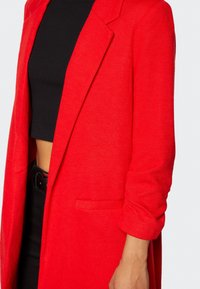 Blazer de mezcla de lana roja con solapas, un bolsillo y mangas enrolladas. La tela presenta un sutil patrón texturizado.