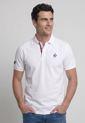 Hombre con polo blanco de manga corta con pequeño logo negro en el pecho, de pie con las manos en los bolsillos, mirando hacia la derecha.