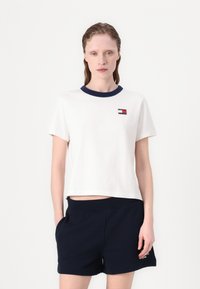 Hvid bomuld T-shirt med navy rund hals, korte ærmer og et lille logo-patch i rød, hvid og blå farver på brystet.