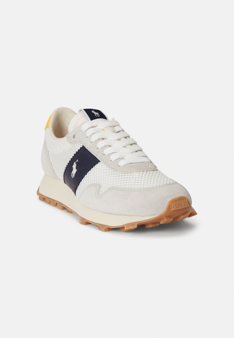 Baskets blanches de sport avec accents navy, matériaux en daim et mesh, semelle en caoutchouc, et marquage visible sur le côté et la languette. Détail jaune sur le talon.