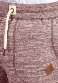 Bordo spalvos sportinės kelnės su struktūruota medžiaga, elastinga juosta, virvutės uždarymu ir rudos odos logotipo pamušalu šone.
