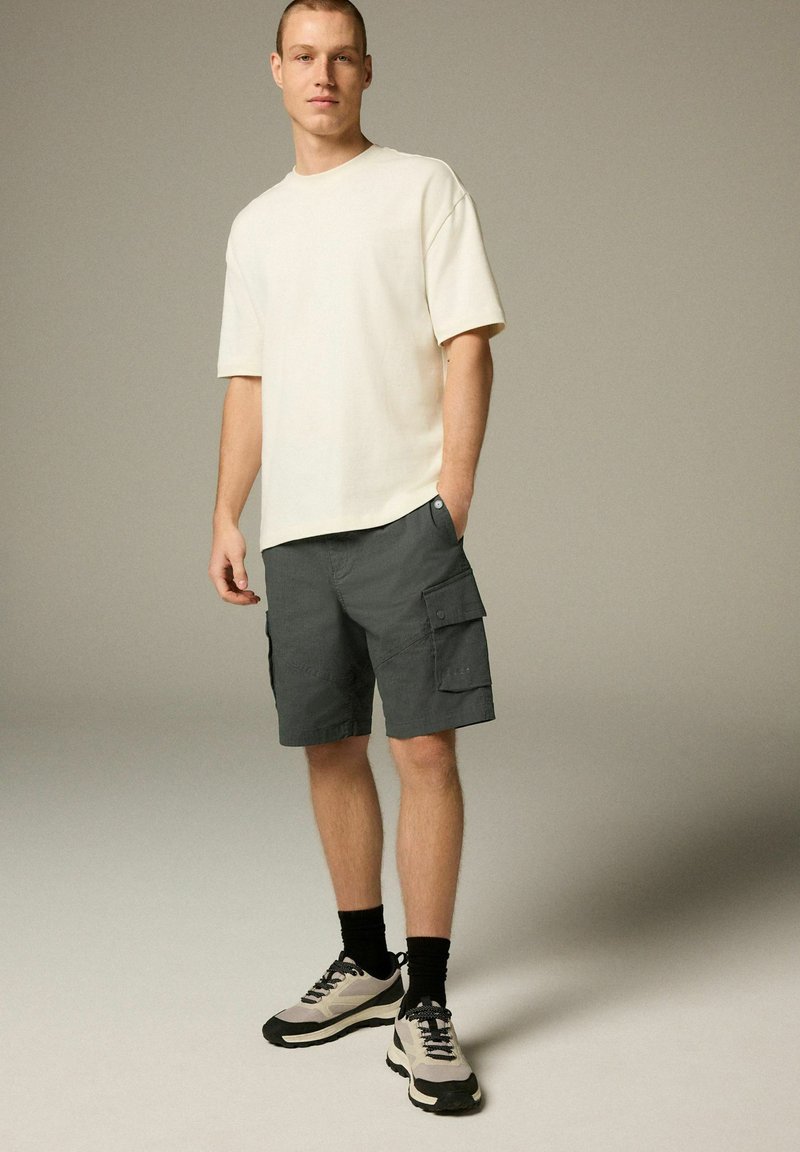 Cremefarbenes Kurzarm-T-Shirt kombiniert mit dunkelgrauen Cargo-Shorts mit Taschen. Das Modell trägt graue und schwarze Sneaker mit dunklen Socken.
