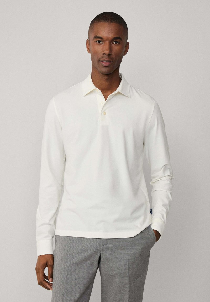 Polo a maniche lunghe, bianco, con colletto classico e tre bottoni; abbinato a pantaloni grigi su misura. Il tessuto appare liscio e leggero.