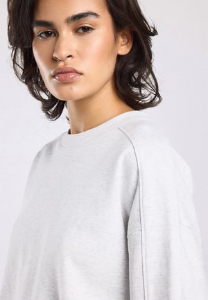 Jong persoon met halflang donker haar, draagt een lichtgrijze sweater met ronde hals, licht zijwaarts gericht tegen een effen achtergrond.