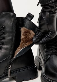 Botas de tobillo de cuero negro con suela resistente, apertura con cremallera y forro de piel de oveja marrón, que muestran un interior texturizado y un acabado exterior suave.