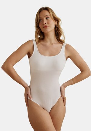 Femme aux cheveux blonds portant un body beige sans manches, les mains sur les hanches, regardant vers sa gauche sur un fond clair uni.