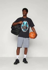 Camiseta gráfica negra con un diseño multicolor en el centro, combinada con pantalones cortos deportivos grises, zapatillas negras y una bolsa negra. Sosteniendo un balón de baloncesto.