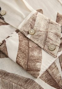 Kreemivärviline lühikeste varrukate shirt, millel on pruun botaaniline lehtede mustriline trükk, kaeluse juures nupud ja sile, kerge tekstuur.